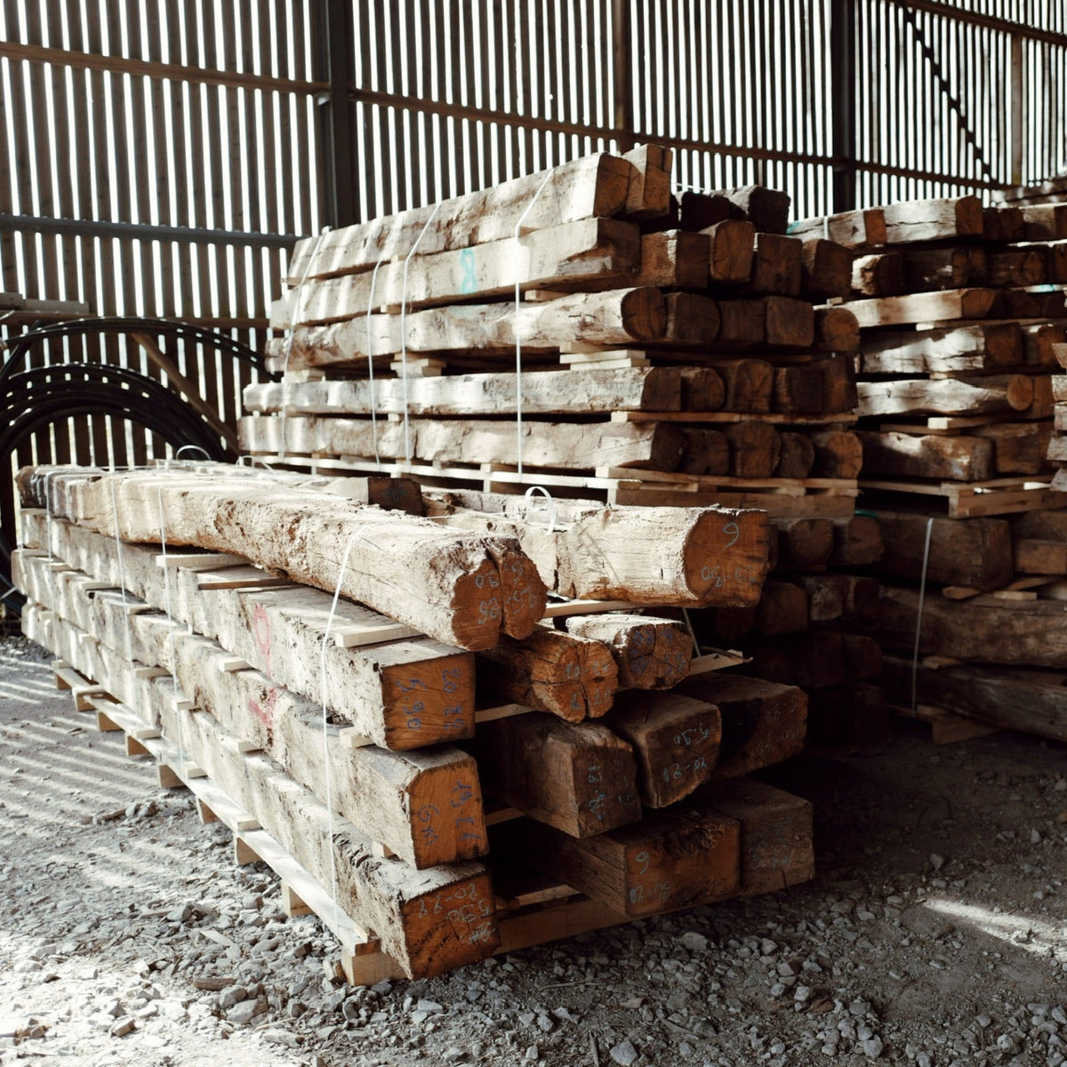 Ancient reclaimed French oak beams stacked in a warehouse, showcasing their weathered textures and unique character.
