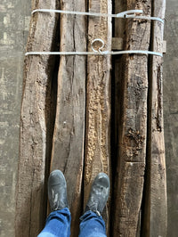 Ancient reclaimed French oak beams showcasing weathered texture and patina, bound together on a floor with person's feet in view.